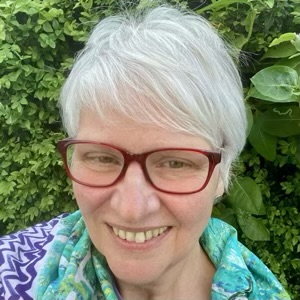 Older white person with short grey hair wearing red freaked glasses and a multicoloured turquoise scarf